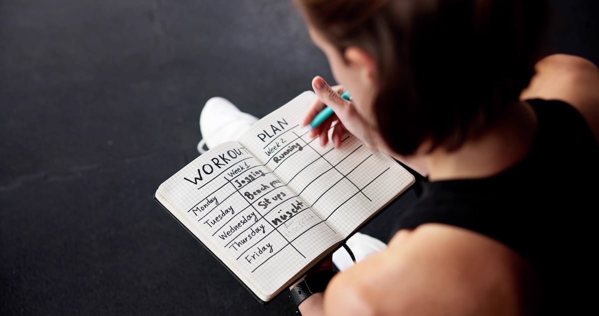 Person writing a weekly workout plan in a notebook, focusing on weight loss routines and exercise goals.