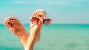 Feet in pink flip-flops with red toenail polish, wearing sunglasses, resting by the beach, symbolizing relaxation during vacation while maintaining weight loss goals.