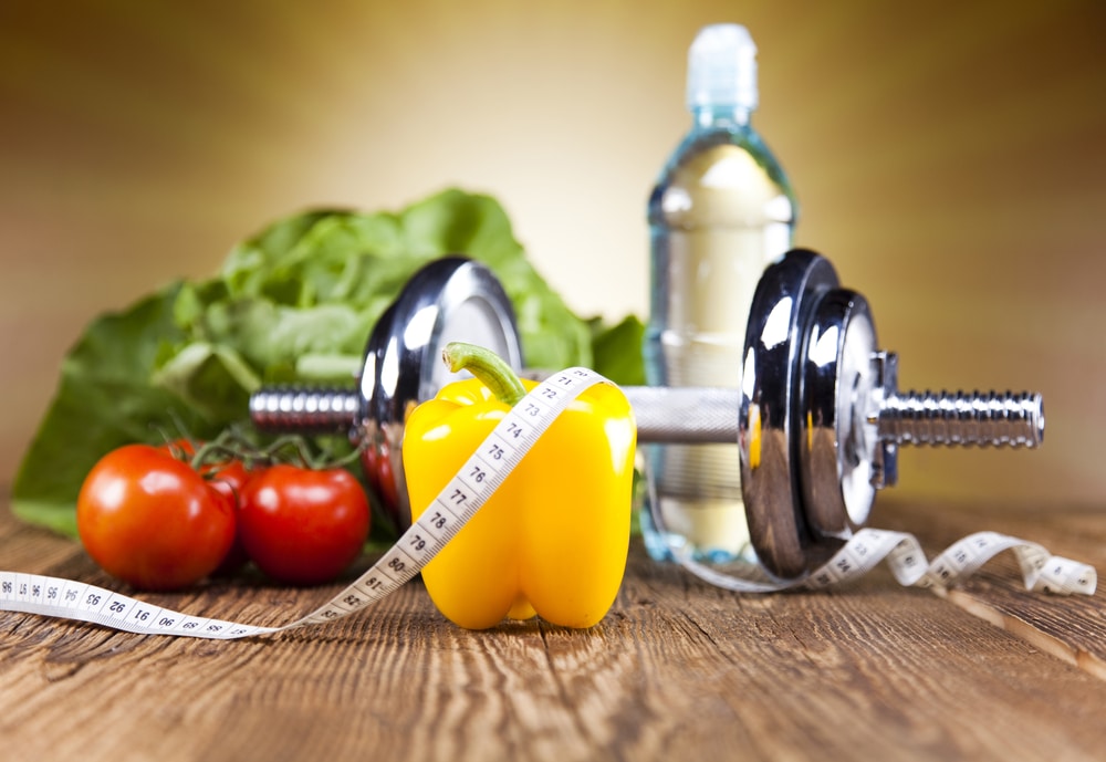 Dumbbell, yellow bell pepper, tomatoes, and measuring tape on wooden surface, symbolizing healthy habits and weight management.
